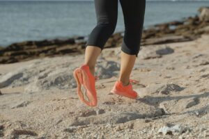 Beach to Bay Relay 2025: Close-up of a runner’s legs wearing bright coral running shoes, jogging along a sandy beach trail with the ocean and rocky shoreline in the background.
