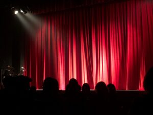 Corpus Christi’s Ritz Theatre: A dimly lit theater stage with closed red curtains, illuminated by a spotlight from the upper left, with silhouettes of audience members seated in the foreground.