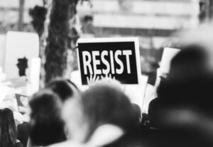 A black and white photo of a crowd at a demonstration, with a bold “RESIST” sign held high, symbolizing the upcoming No Kings Protest Set.