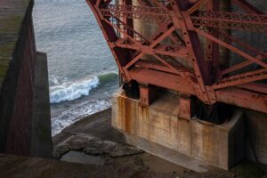 Rust-streaked steel beams anchor a bridge support above crashing waves, symbolizing South Padre’s Bridge of Sorrow