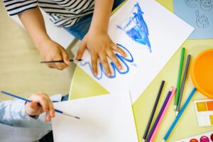 Children paint colorful handprints and drawings with pencils and brushes at a classroom table during a Coastal Bend College activity.