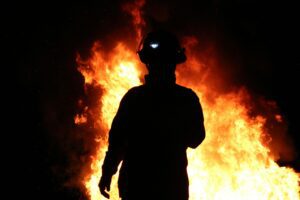 A firefighter stands before blazing flames at night, symbolizing the intense Gregory Cotton Fire Forces battle.
