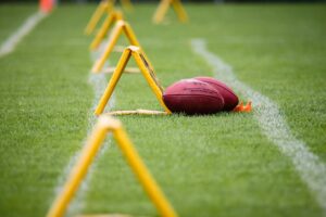 Close-up of footballs on a grassy field beside yellow training hurdles, symbolizing JJ Watt’s Glow-Up from player to analyst.