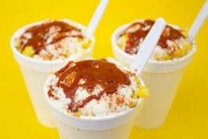 Three cups of elote topped with chili sauce and cheese from The Corpus Christi Food Truck on a bright yellow background.