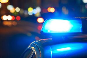 Police vehicle’s blue emergency light flashes at night amid blurred city lights — Man charged after fatal shooting.
