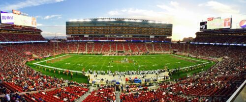Wide view of a packed football stadium at sunset as teams line up on the field, capturing game energy of Shay Whitcomb.