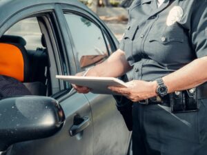 Police officer writes a citation beside a car during a roadside stop, holding a notepad while speaking to the driver.