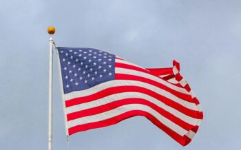 American flag waves against a cloudy sky, symbolizing patriotism and respect for military service. Honoring Veterans Who Stand.