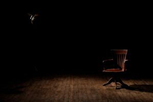 A single wooden chair sits in a dim spotlight on a worn wooden floor, surrounded by darkness.