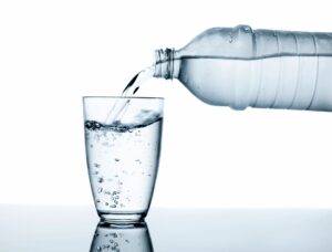 Clear bottled water pours into a glass on a reflective surface, showing bubbles as the glass fills against a white background