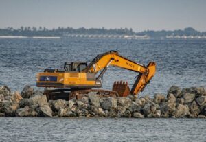 Excavator on rock jetty works along coastal shoreline with ocean in background during marine construction.
