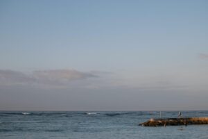 Calm coastal waters at sunset with a small rocky pier, gentle waves, and people fishing and swimming near the shoreline.