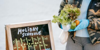 Person holding fresh produce beside a chalkboard displaying a holiday schedule, symbolizing modified services during the winter holidays.