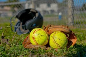 Two softball balls in a worn glove on grassy field with helmet behind fence, Houston Texans practice-themed image