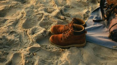 Brown boots resting on sandy beach beside a bag, symbolizing mild winter weather and warm, sunny holiday conditions.