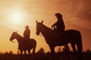 Silhouetted cowboys on horseback pause at sunset, warm golden sky behind them and trees on the horizon.