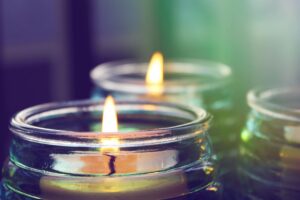 Close-up of lit candles in clear glass jars, soft flames glowing against a blurred green and purple background.