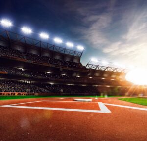 Wide view of a baseball stadium at sunset, with bright lights on, a packed crowd, and home plate centered on the field.