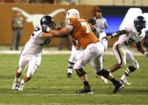 Texas Longhorns offensive lineman blocks a defender during a nighttime college football game, showcasing strength and intensity at the line of scrimmage.