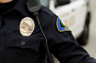 Close-up of a police officer’s uniform showing a badge, shoulder patch, and radio microphone, with a patrol vehicle blurred in the background.