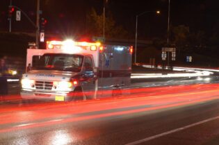 Ambulance with flashing emergency lights stopped at a nighttime intersection as blurred traffic streaks past during an emergency response. spring break crash