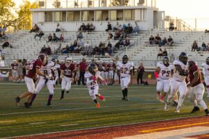 High school football game with players running on field and fans in bleachers, symbolizing coach Leyton Hernandez legacy