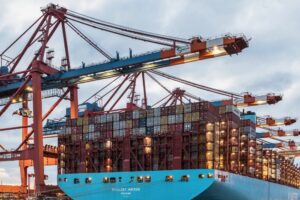 DP World container ship loaded with cargo containers beneath towering cranes at a busy international shipping port.