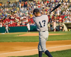 Baseball player swings during a game as fans watch from the stands, illustrating the Frisco RoughRiders in action.